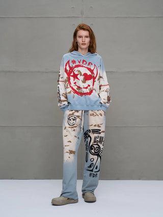 Junge Erwachsene in einem #VDR Pant 18002 blue/beige, mit entspannter Passform, elastischem Bund und abstrakten Grafiken auf dem linken Bein, stehend vor einer grauen Wand.