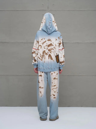 Person trägt #VDR Pant 18002 blue/beige, ein oversized Modell mit gebleachtem Muster und braunen Spritzern, Patch-Detail auf der Kapuzenseite, kombiniert mit schlichten Slip-Ons.