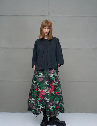 Junge Person trägt #VDR Tee 18007 black, ein oversized schwarzes T-Shirt mit subtil geprägtem Frontprint, kombiniert mit einer voluminösen Camouflage-Rock und klobigen schwarzen Stiefeln vor grauer Wand.