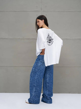 Junge Frau in Profilansicht trägt das #VDR Tee 18037 white: Ein oversized, asymmetrisches T-Shirt mit rosenförmigem Rückenprint, kombiniert mit hochgeschnittenen, weiten Jeans und weißen Sneakers.