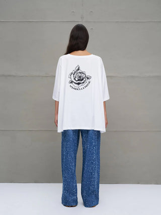 Person mit dem Rücken zur Kamera trägt das #VDR Tee 18037 white: Oversized weißes T-Shirt mit großem Rosen-Print und VIADELLEROSE-Schriftzug auf der Rückseite, kombiniert mit blauen, lockeren Hosen.