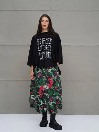 Junge Frau trägt #VDR Tee 18042 black, ein locker sitzendes, schwarzes, dreiviertelärmliges Oberteil mit weißem Slogan-Aufdruck, kombiniert mit voluminösem Camouflage-Rosen-Rock und klobigen schwarzen Plateaustiefeln.