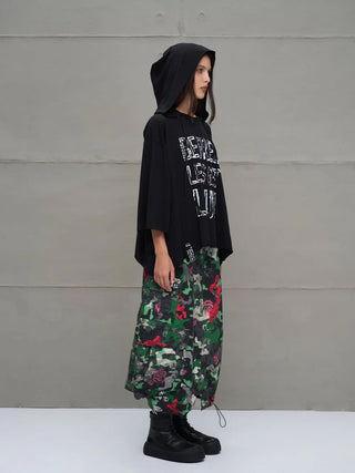 Junge Frau im Profil trägt das #VDR Tee 18042 black, einen übergroßen schwarzen Kapuzenpullover mit asymmetrischem Schnitt und auffälligem Slogan-Frontprint, kombiniert mit einer langen Camouflage-Rosen-Rock.