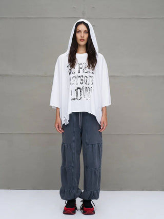Person trägt #VDR Tee 18042 white, ein oversized, weißes Kapuzen-T-Shirt von Viadellerose mit asymmetrischem Schnitt und Slogan-Print, kombiniert mit lässigen, gestuften, grauen Hosen und roten Sneakers.