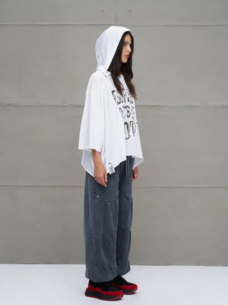Eine junge Frau trägt das #VDR Tee 18042 white, ein oversized, weißes, asymmetrisches Kapuzen-T-Shirt mit schwarzem Slogan-Print und Nieten-Details, kombiniert mit weiten, grauen Hosen.