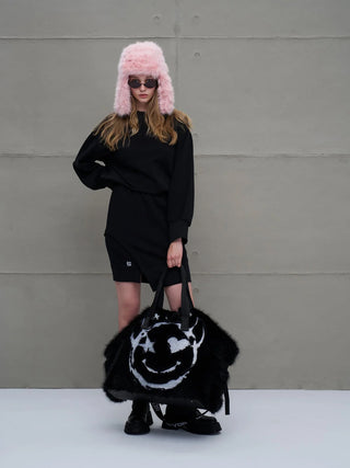 Modell trägt den #VDR Pan Hat pink, eine flauschige, pastellrosa Mütze mit Ohrenschützern und Nackenverschluss, kombiniert mit einem schwarzen Outfit und einer übergroßen, schwarzen Tasche.