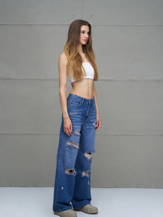 Junge Frau in #VDR Destroy Denim 18132 midblue, hochgeschnittene, weit geschnittene Jeans mit markanten Rissen, trägt ein weißes, trägerloses Oberteil, entspannt posierend gegen eine graue Wand.
