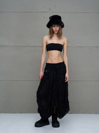 Junge Frau trägt #VDR Rock 18170 black, präsentiert als fließender Midi-Rock mit fransigen Details und asymmetrischem Saum, kombiniert mit einem schwarzen Bandeau-Top und Plattformsandalen.