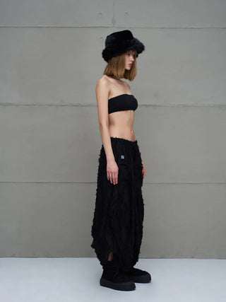 Seitenansicht einer Frau, die den #VDR Rock 18170 black trägt: ein schwarzer, semi-transparenter Midi-Rock mit asymmetrischem Saum und fransigen Details, kombiniert mit einem schwarzen Bandeau-Top.