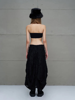 #VDR Rock 18170 black: Person in schwarzem, halbtransparentem Midi-Rock mit asymmetrischem Saum und fransigen Details, kombiniert mit schwarzem Bandeau-Top und klobigen Plattformen, von hinten vor grauer Wand stehend.
