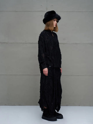 Person in Profilansicht trägt das #VDR Shirt 18171 black, ein locker sitzendes, semi-transparentes Oberteil mit fransigen Details und Mandarinkragen, kombiniert mit weiten, strukturierten Hosen und einem schwarzen Hut.