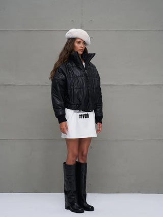 Junge Frau trägt #VDR Jacket 18246 black, eine schwarze, gesteppte Bomberjacke mit Reißverschluss und Stehkragen, über einem weißen Rock. Die Jacke betont sportliche Eleganz und passt perfekt zu urbanen Looks.