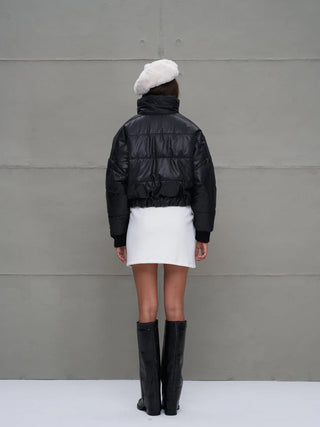 Person von hinten in #VDR Jacket 18246 black, einer kurzen, gepolsterten Bomberjacke mit Stehkragen und Reißverschluss, kombiniert mit weißem Minirock, schwarzen Stiefeln und weißem Beret vor grauer Wand.