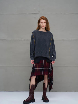 Junge Frau trägt #VDR Sweater 18005 anthrazit, oversized mit diagonalen goldfarbenen Zippern. Darunter schaut eine schwarze Lage hervor. Kombiniert mit einem asymmetrischen Karo-Rock und burgunderroten Stiefeln.