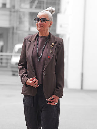 Ältere Frau trägt den OFF#DLY Blazer Fiora braun mit dekorativen Broschen; kombiniert mit schwarzer Sonnenbrille, plumfarbenem Pullover und schwarzen Cargo-Hosen für einen stilvollen urbanen Look.