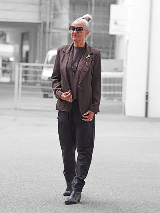 Ältere Frau trägt den OFF#DLY Blazer Fiora braun mit dekorativen Broschen über einem dunklen Pullover, kombiniert mit schwarzen Cargo-Hosen und Stiefeletten, vor einem städtischen Hintergrund.