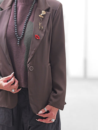 Nahaufnahme des OFF#DLY Blazer Fiora braun mit einem schmalen Schnitt. Verziert mit drei Broschen am Revers: goldene Rose, Hirschumriss und rote Lippen. Getragen über einem burgunderfarbenen Pullover.