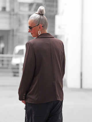 Person in urbaner Umgebung trägt den OFF#DLY Blazer Fiora braun mit lockerem Schnitt, sichtbaren Ärmelnähten und geometrischen Ohrringen, ergänzt durch klassische Sonnenbrille für einen stilvollen Look.