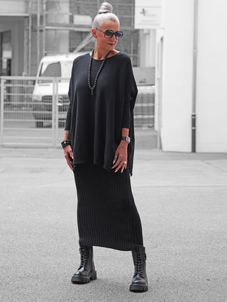 Frau in urbanem Umfeld trägt den OFF#DLY Rock Linda black, einen gerippten Midi-Rock mit elastischem Bund, kombiniert mit einem oversized schwarzen Pullover und klobigen Stiefeln.