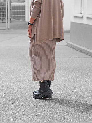 Person trägt OFF#DLY Rock Linda taupe, ein geripptes Midi-Design mit elastischem Bund, kombiniert mit schwarzen, klobigen Stiefeln und einem lockeren Strickoberteil, sichtbar in urbaner Umgebung.