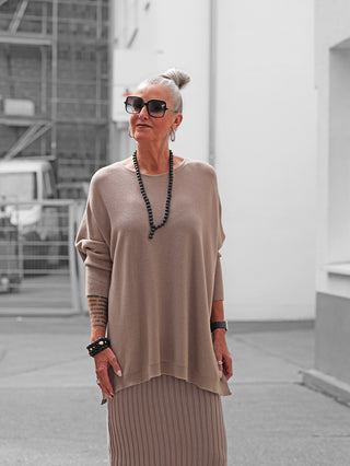 Ältere Frau in taupefarbenem OFF#DLY Pulli Olivia, oversize, mit weicher Textur, kombiniert mit passenden Strickrock. Accessoires: schwarze Sonnenbrille, lange schwarze Perlenkette, Armreifen, Ringe. Ruhige Stadtstraße im Hintergrund.