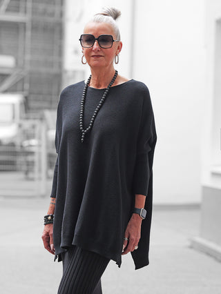 Ältere Frau trägt den OFF#DLY Pulli Olivia schwarz mit Oversize-Schnitt, langen Ärmeln und seitlichen Schlitzen, kombiniert mit schwarzen Leggings und Accessoires, vermittelt lässigen, eleganten Look.