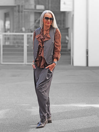 Reife Frau trägt OFF#DLY Baggy Pant Abby anthrazit, locker geschnittene graue Hose mit leicht zulaufendem Bein, kombiniert mit rustbrauner Bluse und grauem Vest, Hände in Hosentaschen.
