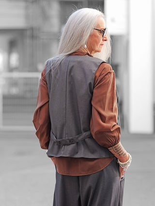 Ältere Frau im urbanen Umfeld trägt die OFF#DLY Weste Alison grey, die durch schlichte Eleganz besticht, über einer rostbraunen Bluse, mit Perlenarmband und blockförmigem Tattoo am Unterarm.
