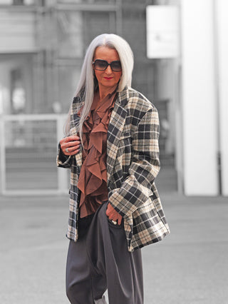 Eine stilvolle Frau trägt den Lumina Blazer black/white mit klassischem Karomuster und Reverskragen, kombiniert mit einer braunen Bluse. Der Blazer hat gepolsterte Schultern und Pattentaschen.