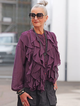 Ältere Frau trägt die OFF#DLY Bluse Rita bordeaux mit rüschigem Design und luftiger Passform, kombiniert mit dunklen Hosen und Accessoires; ideal für einen eleganten, femininen Look.