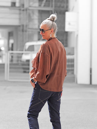 Ältere Person in urbaner Umgebung trägt die OFF#DLY Bluse Rita braun, locker geschnitten mit verspielten Volants, kombiniert mit dunklen Jeans, Sonnenbrille, Smartwatch, großen Ohrringen und Halskette.