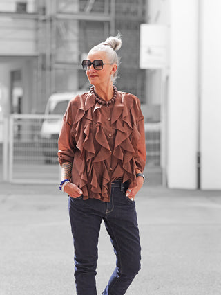 Eine Frau trägt die OFF#DLY Bluse Rita braun mit verspielten Volants und lockerem Schnitt. Kombiniert mit Jeans und Accessoires, zeigt sie einen stilvollen, urbanen Look.