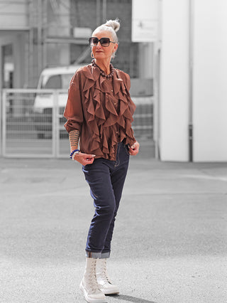 Person trägt die OFF#DLY Bluse Rita braun mit auffälligem Volant-Design und lockerer Passform, kombiniert mit dunklen Jeans und weißen Stiefeln. Accessoires: Halskette, Ohrringe, Armbänder.