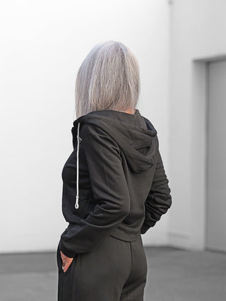 Eine Person trägt die OFF#DLY Sweatjacke Sulden II black, eine schmal geschnittene, schwarze Kapuzenjacke mit Reißverschluss. Die Hände sind in den seitlichen Taschen verstaut.
