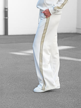 Person trägt OFF#DLY Sweatpant Stilfs white: weiße, locker geschnittene Jogginghose mit dunklen Streifen entlang der Seiten, kombiniert mit weißen Sneakers, Hand in Tasche, sichtbare Ringe.