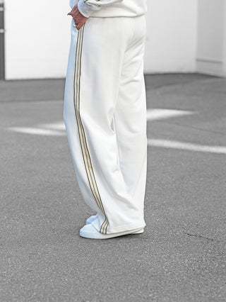 Person trägt OFF#DLY Sweatpant Stilfs white: lockere weiße Trackpants mit goldenen Seitenstreifen, Hand in Tasche, kombiniert mit weißen Sneakers.