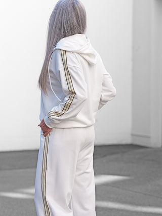 Person von hinten in der OFF#DLY Sweatjacke Sulden white, eine Kapuzenjacke mit Kontraststreifen an den Ärmeln. Sie trägt dazu passende, weit geschnittene Jogginghosen mit ähnlichen Streifendetails.