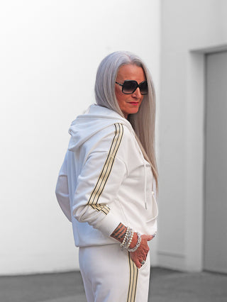 Eine Frau in der OFF#DLY Sweatjacke Sulden weiß mit goldenen Streifen, Kapuze, Reißverschluss und Taschen, lässig posierend mit einem Tattoo am Arm und Sonnenbrille.