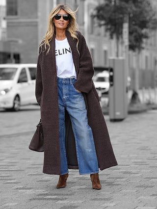 Frau trägt Gitta Banko MEI Mantel Marone: langer, gerade geschnittener Teddy-Mantel, kombiniert mit weißem Logo-T-Shirt, blauer Jeans, braunen Stiefeln und Tasche, perfekt für stilvolle, komfortable Stadtausflüge.