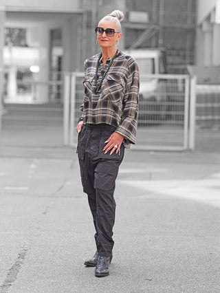 Ältere Frau trägt die Nuomeno Bluse Raglan brown check mit feinem Karomuster und Raglan-Ärmeln, kombiniert mit schwarzen Cargo-Hosen und Stiefeln in urbaner Umgebung.