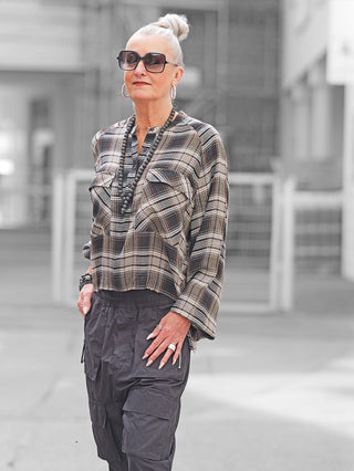 Ältere Frau in urbanem Umfeld trägt Nuomeno Bluse Raglan brown check mit lockerem, kariertem Muster und Raglan-Ärmeln, kombiniert mit schwarzen Cargo-Hosen und auffälligen Accessoires.