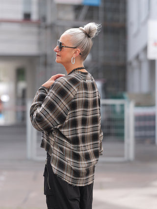 Eine Frau trägt die Nuomeno Bluse Raglan brown check mit Raglan-Ärmeln und elastischen Abschlüssen, ideal kombiniert mit schwarzen Cargo-Hosen für einen urbanen, lässigen Look.