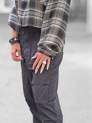 Person trägt die Nuomeno Bluse Raglan brown check, eine karierte Raglan-Ärmelbluse, in dunklen Cargo-Hosen. Hände zeigen chunky Ringe und beaded Armbänder.