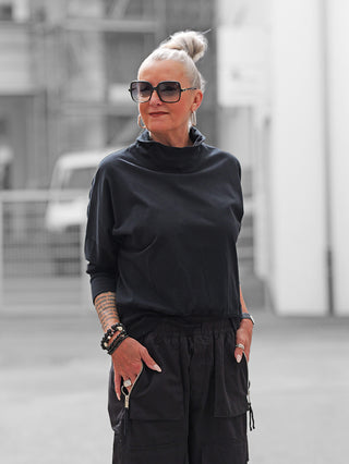 Frau trägt Nuomeno Tee Dolcevita nero; schwarzes, hochgeschlossenes Langarmshirt mit markantem Kragen. Kombiniert mit schwarzen Cargo-Hosen und Schmuck, zeigt es modernes, minimalistisches Design.