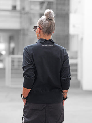 Eine Person von hinten mit dem Nuomeno Tee Dolcevita nero, erkennbar am markanten Kragen und den langen Ärmeln, kombiniert mit lockeren Cargo-Hosen in einer urbanen Umgebung.