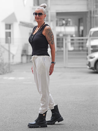 Ältere Frau in Nuomeno Pant offwhite, entspannt stehend mit schwarzem Tanktop und klobigen Stiefeln, zeigt die schmal geschnittene Jogginghose mit elastischem Bund und praktischen Taschen.