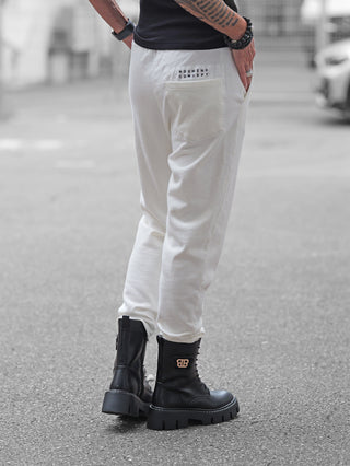 Person trägt Nuomeno Pant offwhite, eine schmal geschnittene Jogginghose mit elastischem Bund und Logo-Print hinten, kombiniert mit schwarzen Combat Boots und dezentem Schmuck.