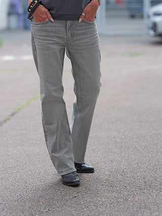 Eine Person trägt True Religion KATE Straight Jeans in Grau mit geradem Bein, kombiniert mit schwarzen Schnürschuhen, auf einem asphaltierten Untergrund. Hände in den Taschen, Schmuck sichtbar.