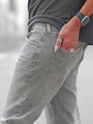 Nahaufnahme der True Religion KATE Straight Jeans in Grau mit dezenter Waschung. Eine Hand mit Ringen steckt im Gesäßtasche. Sichtbare Details: klassische Passform, ikonische Taschenstickerei, komfortabler Stretchanteil.