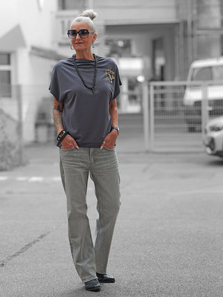 Ältere Frau in True Religion KATE Straight Jeans mit geradem Bein, lockerem grauen Top und Accessoires, steht lässig auf städtischem Gehweg, Hände in den Taschen, Tattoos sichtbar.